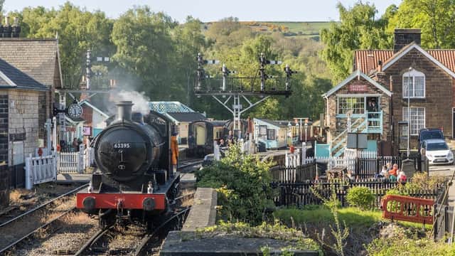 Steam train arriving at Grosmont station, surrounded by lush greenery and quaint buildings, with people enjoying the scenery.