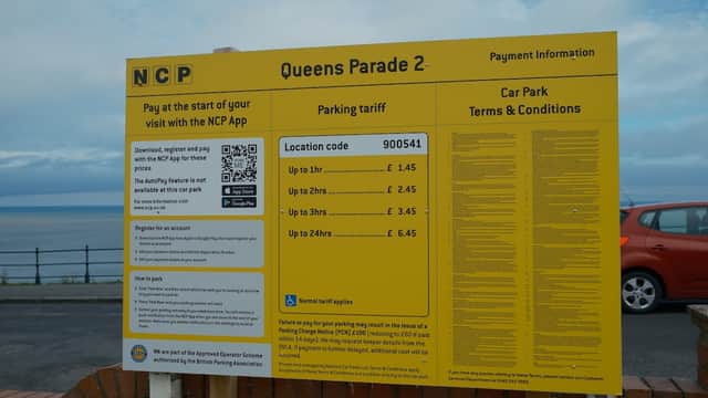 Bright yellow sign displaying parking information and tariffs for NCP Queens Parade 2, with a coastal view in the background.