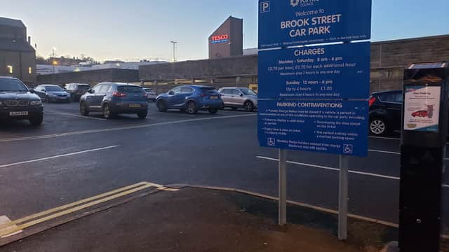 Brook Street Car Park sign with parking charges, surrounded by parked cars and a Tesco store in the background.