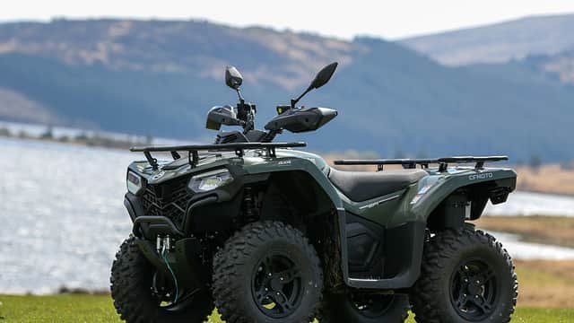 A green utility vehicle parked on grass near a lake, with hills and trees in the background.
