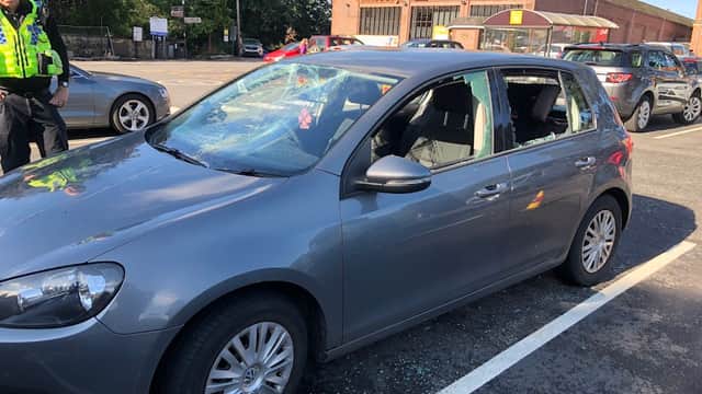 A gray car with shattered windows parked in a lot, with a police officer nearby and shops in the background.