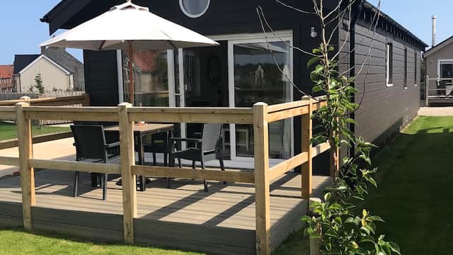 Black wooden cabin with a deck, patio furniture, and an umbrella, set in a green lawn under a clear blue sky.