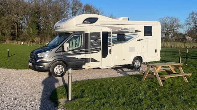 A camper van parked on gravel, surrounded by green grass and trees under a clear blue sky at Laurels Park Campsite.