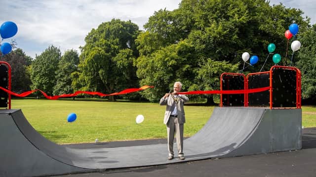 A man in a suit cuts a red ribbon at a skate park, surrounded by trees and balloons in a grassy area.