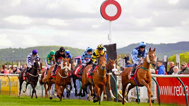 Horses racing at Redcar Racecourse, with colorful jockeys and a large red and white marker in the background.