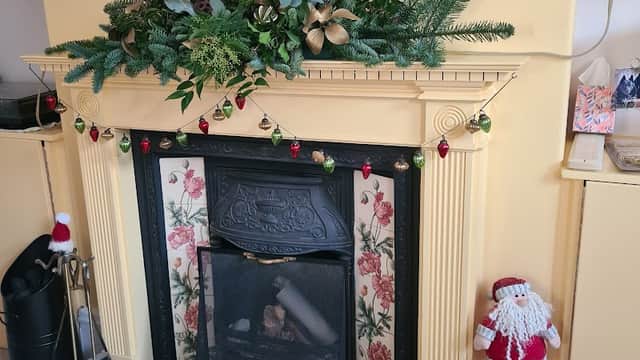 Festive mantel decorated with greenery, ornaments, and a Santa figure beside a black fireplace and wood basket.