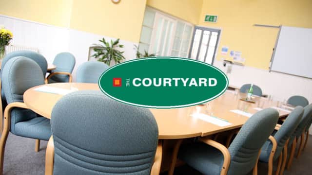 Round table surrounded by green chairs in a bright meeting room with plants and a whiteboard.