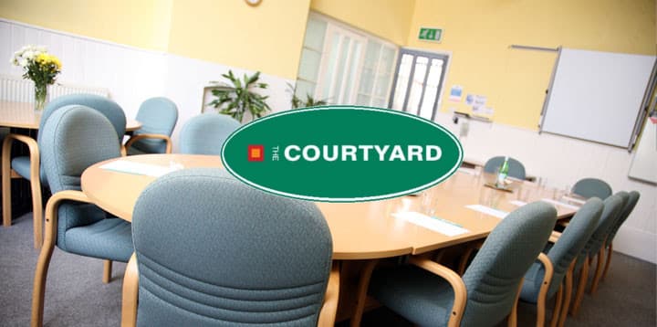 Round table surrounded by green chairs in a bright meeting room with plants and a whiteboard.