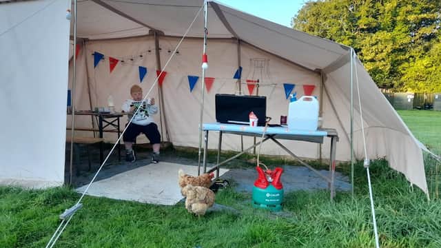 A canvas tent with bunting, a table with supplies, and a person sitting, alongside a chicken on the grass.