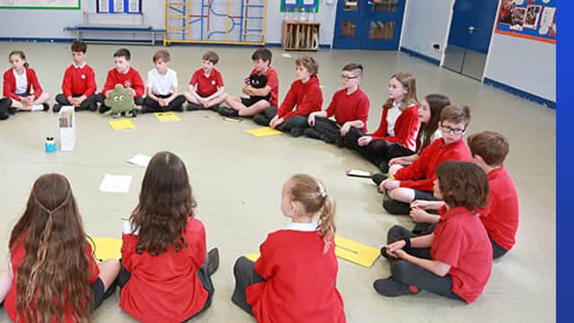 Students in red uniforms sitting in a circle on the floor, engaged in a group activity in a classroom setting.