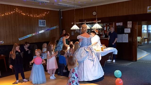 A festive event at Barlow Village Club with children dancing, balloons, and a woman in a blue gown interacting with guests.
