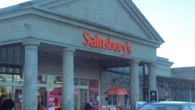 Sainsbury's storefront with columns, featuring Argos signage, shopping carts, and a parked car outside.
