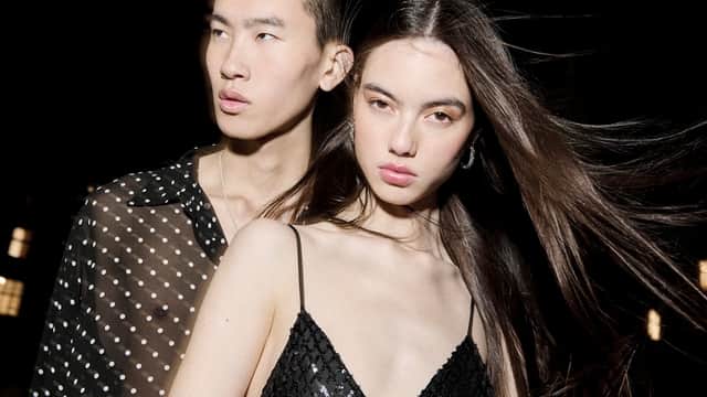 Two models pose closely together, showcasing stylish, modern clothing against a dark background.