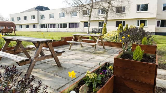 Outdoor seating area with wooden picnic tables, flower beds, and a modern hotel building in the background.