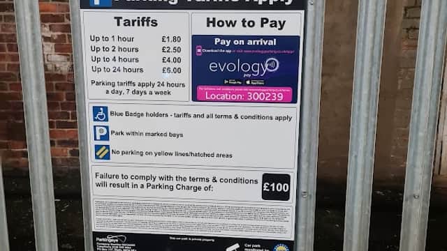 Parking tariff sign with payment instructions and rates displayed on a metal fence at a car park in Middlesbrough.