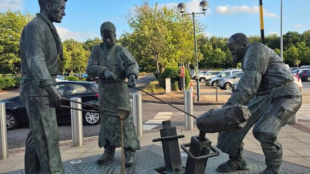 Bronze statue of three workers in a car park, surrounded by trees and parked cars under a blue sky.