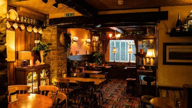 Cozy bar interior with wooden beams, tables, chairs, and warm lighting, featuring a stone fireplace and decorative shelves.