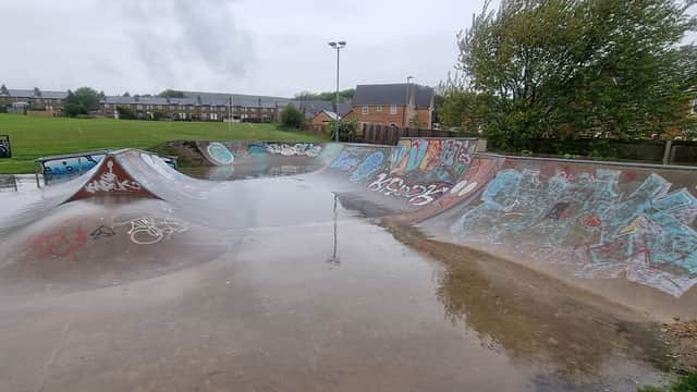 Skatepark with wet concrete ramps, colorful graffiti, and grass fields in the background under a cloudy sky.