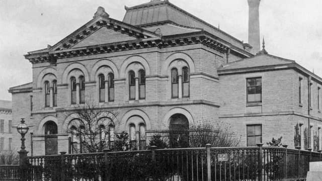 Historic building with arched windows, decorative stonework, and a prominent chimney, surrounded by greenery and a fence.