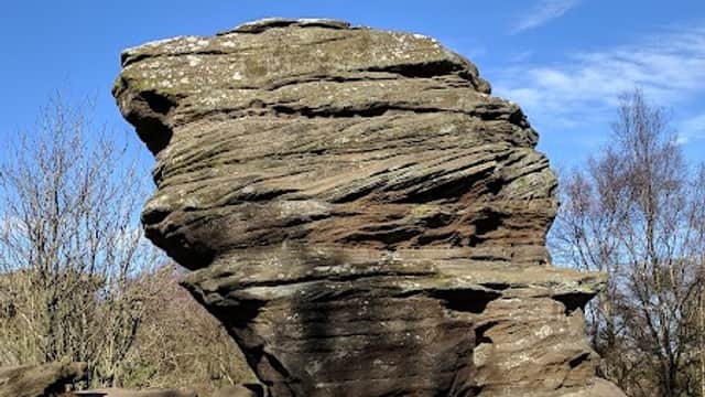 Large, weathered rock formation with layered textures, surrounded by grass and sparse trees under a clear blue sky.