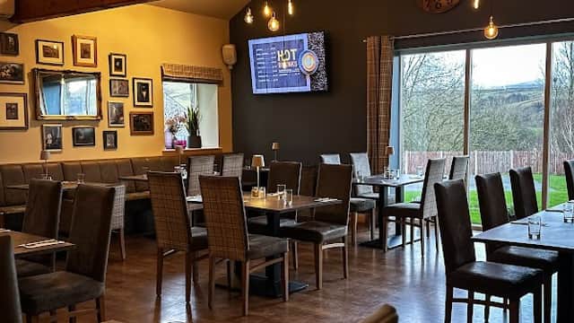 Rustic dining area with wooden beams, stylish chairs, and large windows overlooking greenery. Wall adorned with framed photos.