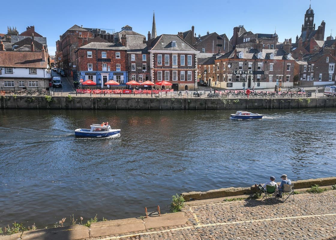 Queens Hotel along the river in York, with boats passing and outdoor seating under red umbrellas.