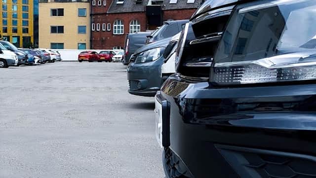 Cars parked in a spacious lot with modern buildings and a cloudy sky in the background.