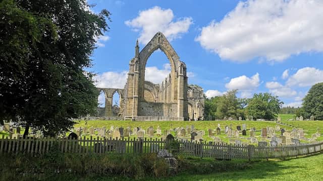Ruins of Bolton Abbey with a grassy area and gravestones, framed by trees under a blue sky with fluffy clouds.
