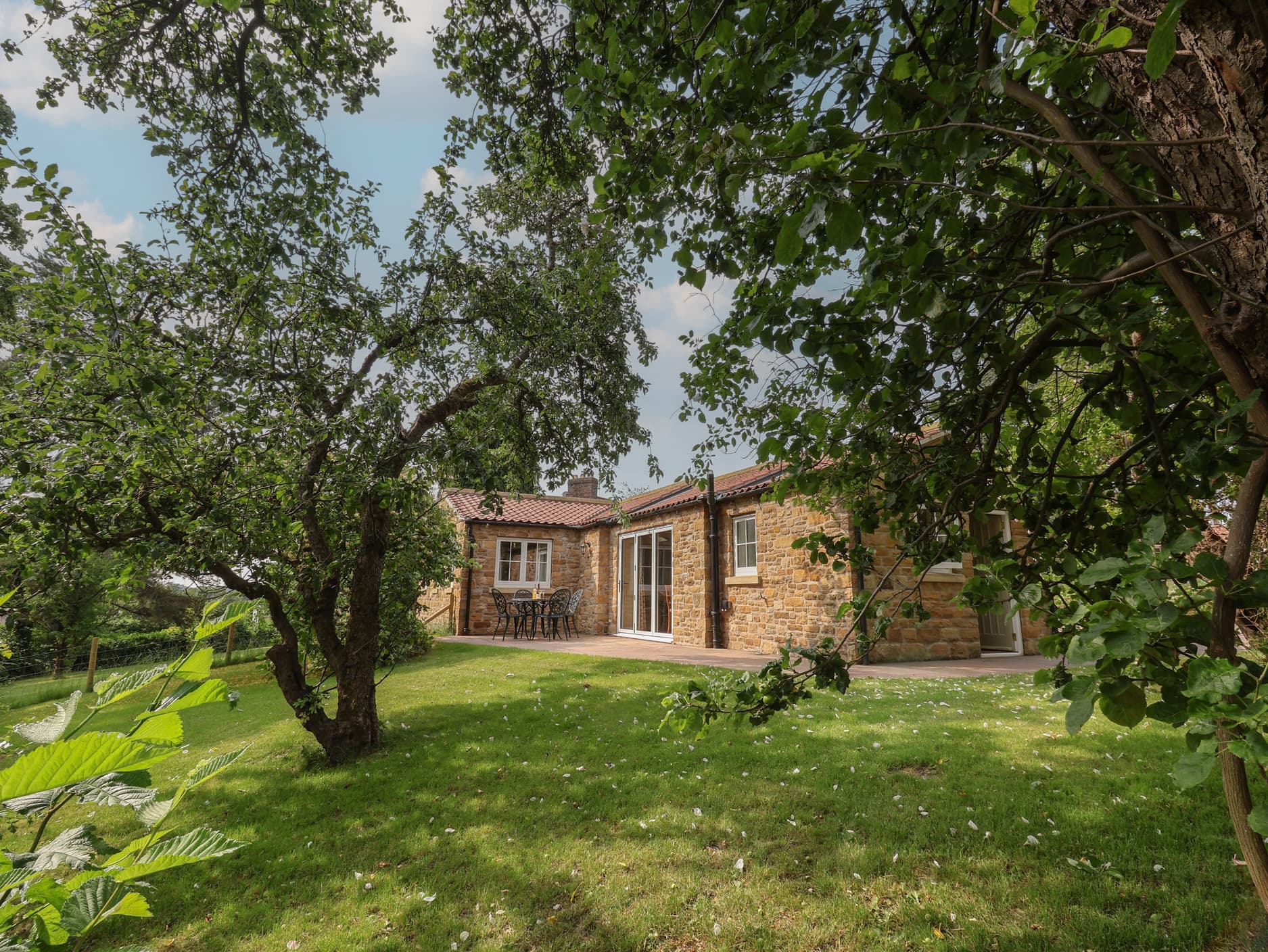 Stone cottage with red-tiled roof, surrounded by lush greenery and apple trees, set on a grassy lawn.