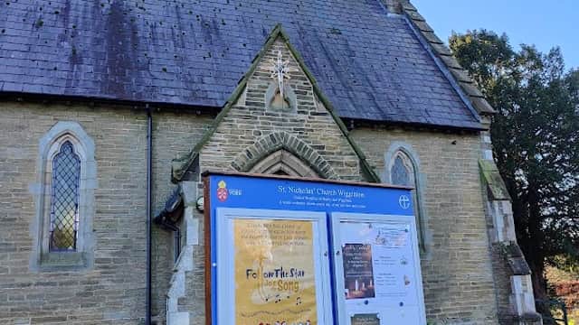 St Nicholas Church with a stone facade, a prominent steeple, and an information board in front against a clear blue sky.