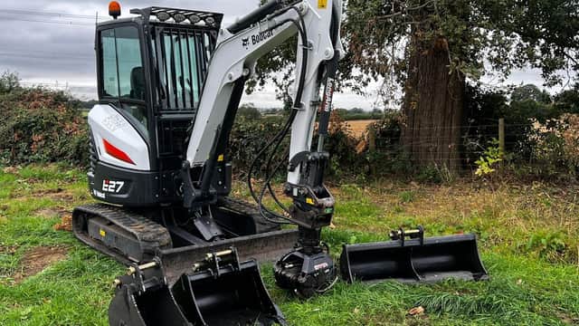 Excavator parked on grass with two attachments, surrounded by trees and open fields in North Yorkshire.