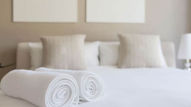 Neatly rolled white towels on a crisp bed with soft pillows against a neutral wall.