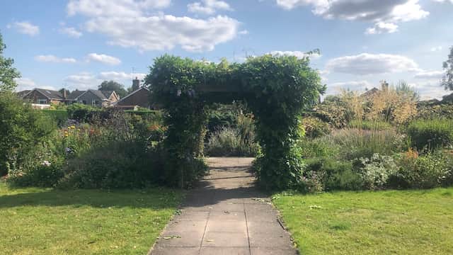 A lush garden path leads through a green archway, surrounded by colorful flowers and under a bright blue sky.