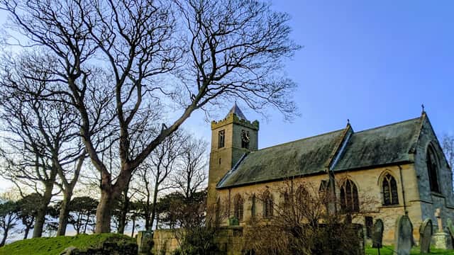 All Saints Church with a tall tree nearby, set against a clear blue sky and grassy grounds.