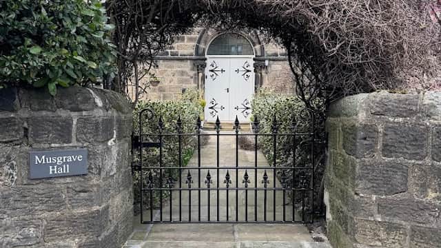 Archway covered in vines leads to a white door at Musgrave Hall, framed by stone walls and a paved pathway.