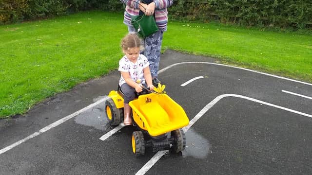 A child rides a yellow toy tractor on a play area path while an adult watches nearby in a green field.