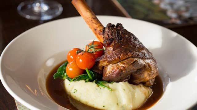 A plated dish featuring a lamb shank, cherry tomatoes, spinach, and creamy mashed potatoes in rich gravy.