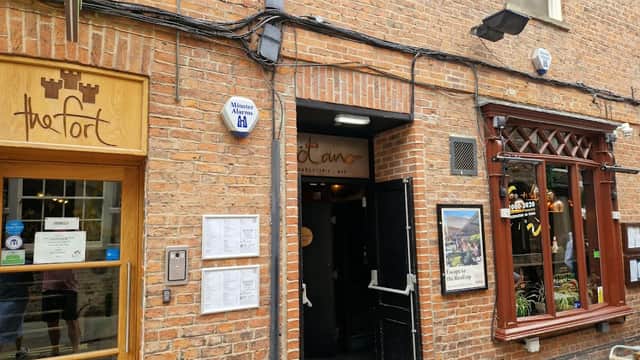 Entrance to Manahatta York pub, featuring a brick facade and wooden accents, with signage and outdoor seating visible.