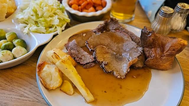 A plate of roast beef with gravy, Yorkshire pudding, potatoes, carrots, cabbage, and a pint of beer on a wooden table.