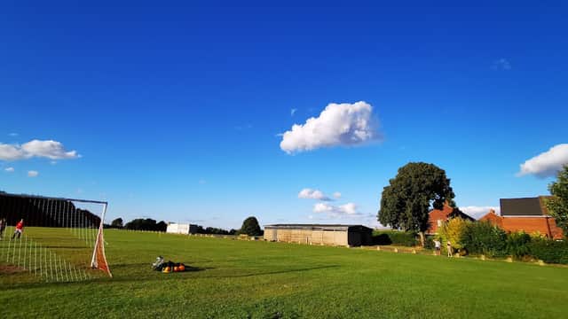 Clear blue sky over a green field with a soccer goal, surrounded by trees and buildings in the distance.