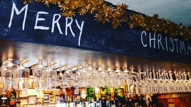 Bar interior with festive decorations, "MERRY CHRISTMAS" written on a chalkboard, and rows of hanging glasses.