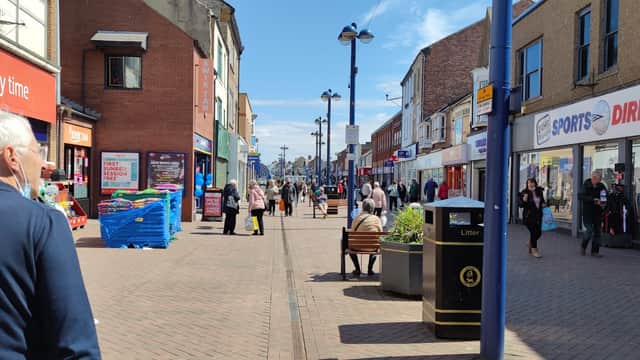 Busy street scene in Redcar with shops, pedestrians, and clear blue skies. Benches and a bus stop are visible.