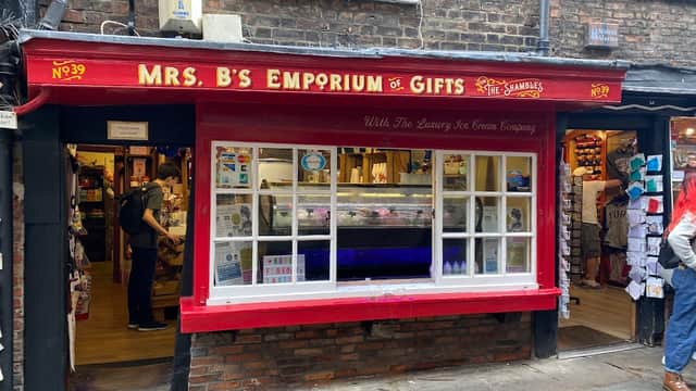 Red storefront of Mrs B's Emporium of Gifts with large window displaying gifts and ice cream, cobbled street outside.