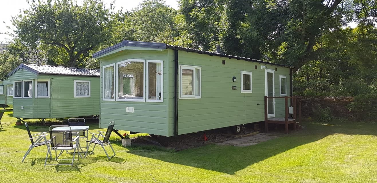 Green caravan with large windows, surrounded by grass and trees, at Orchard Caravan & Camping Park in Reeth, Yorkshire.