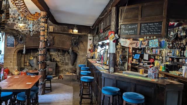Rustic bar interior with wooden beams, a stone fireplace, colorful stools, and shelves stocked with various drinks.