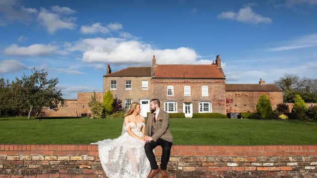 A couple in wedding attire sits on a brick wall in front of Hornington Manor, surrounded by green lawns and blue skies.