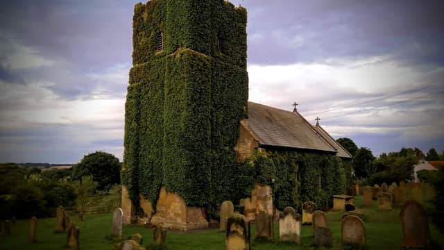 St. John The Evangelist Church covered in ivy, surrounded by gravestones and lush greenery under a cloudy sky.