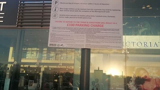Sign for Blue Badge holders only, detailing parking rules and potential charges, with a shopping center in the background.