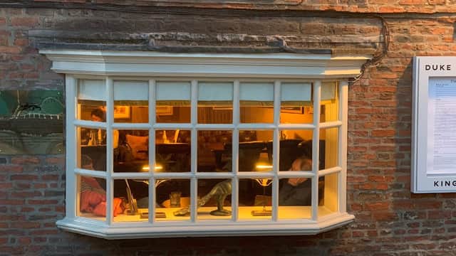 Cozy pub window with warm lighting, brick wall exterior, and patrons enjoying drinks inside.