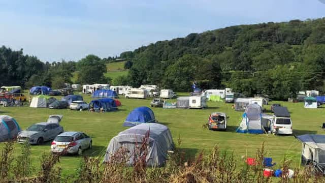 Camping site with numerous tents and caravans set in a green landscape, surrounded by trees under a clear blue sky.